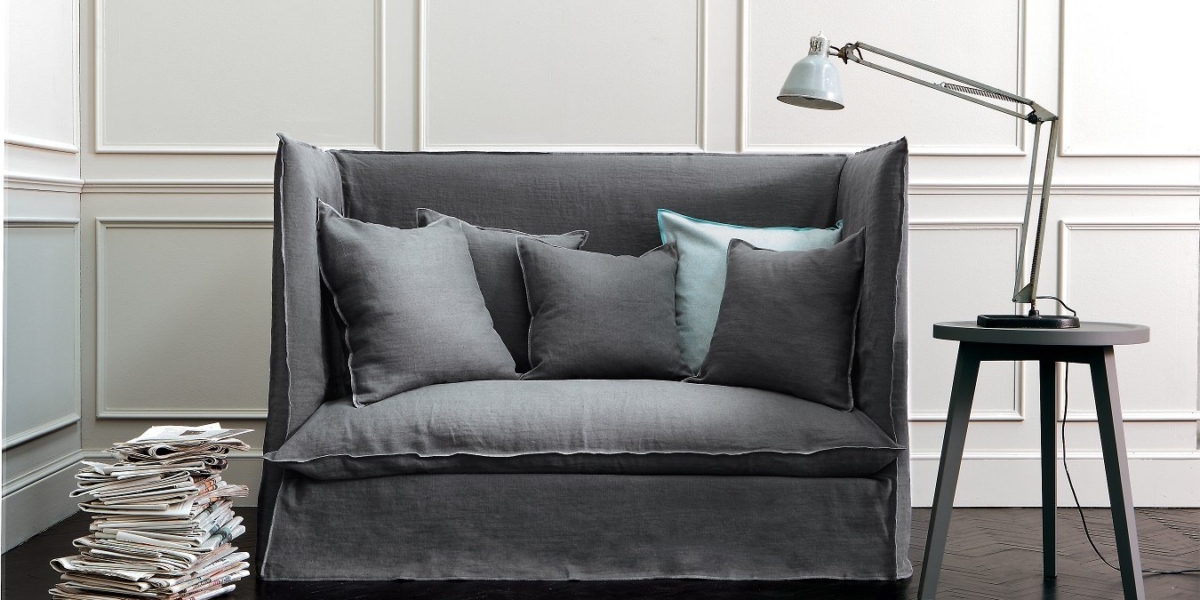 Un canapé gris à dossier haut avec des coussins, une table d'appoint avec une lampe et un tapis bleu s'associent parfaitement dans cette pièce moderne d'inspiration parisienne. Une pile de magazines ajoute une touche de qualité, la rendant à la fois élégante et confortable. Canape Paris by Atelier Bis