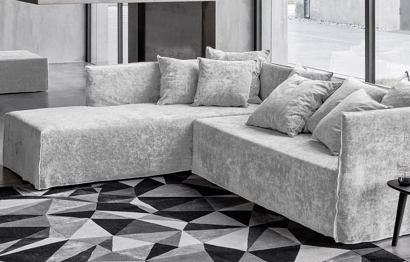 Un salon moderne digne d'une salle d'exposition comprend un canapé gris en forme de L, complété par un tapis géométrique et une petite table d'appoint noire contenant un verre d'eau - un véritable témoignage de style et de qualité. Canape Paris by Atelier Bis