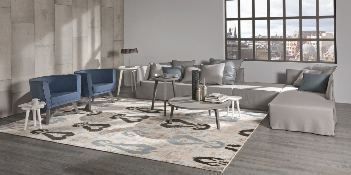 Salon moderne avec canapé d'angle gris, fauteuils bleus et tapis à motifs. Des tables basses sont disposées avec élégance sous la vue sur la ville qui s'étend à travers de grandes fenêtres, donnant à l'espace une qualité de salle d'exposition. Canape Paris by Atelier Bis