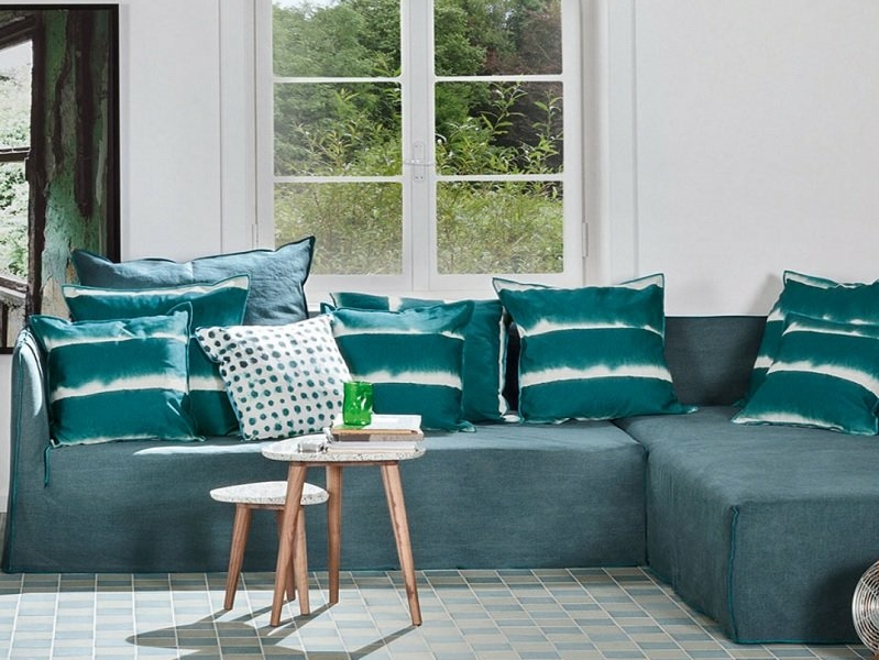 Le salon est doté d'un luxueux canapé sectionnel bleu sarcelle, orné de coussins assortis. Une élégante table d'appoint en bois complète l'aménagement, tandis qu'une grande fenêtre encadre une végétation luxuriante à l'extérieur, rappelant un showroom parisien chic. Canape Paris by Atelier Bis