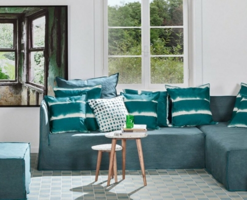 Salon parisien moderne avec un canapé bleu canard, des coussins rayés et une table ronde en bois. Une grande fenêtre met en valeur une végétation luxuriante, ajoutant une touche de qualité naturelle à cet espace chic. Canape Paris by Atelier Bis