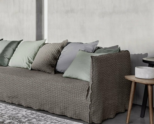 Un canapé gris texturé orné de plusieurs coussins gris et verts trône élégamment à côté d'une petite table d'appoint sur un sol gris chic, rappelant un showroom parisien élégant. Canape Paris by Atelier Bis