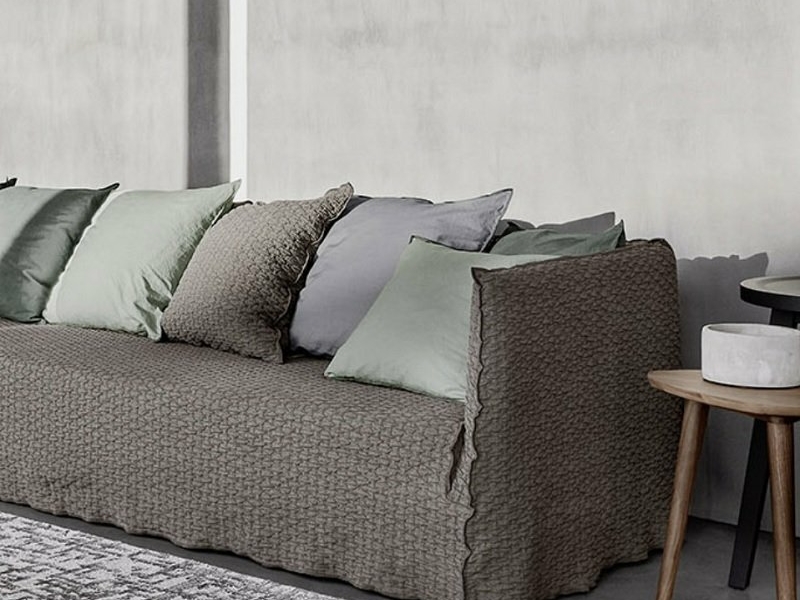 Un canapé gris texturé orné de plusieurs coussins gris et verts trône élégamment à côté d'une petite table d'appoint sur un sol gris chic, rappelant un showroom parisien élégant. Canape Paris by Atelier Bis