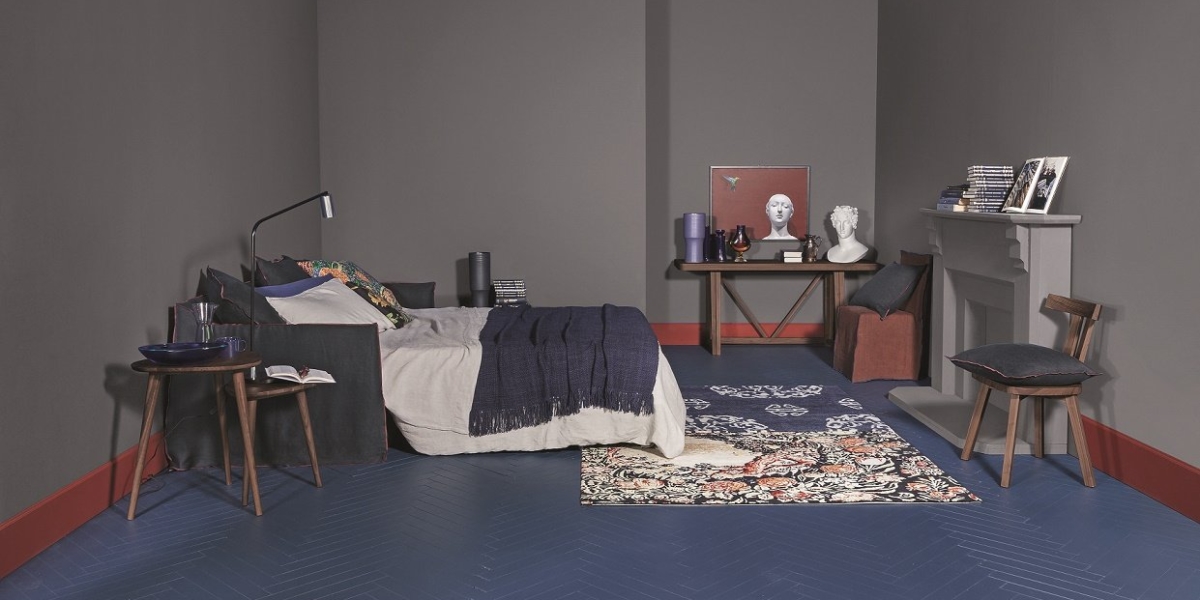 Un showroom sombre et moderne abrite une chambre chic avec un lit douillet, des tables de nuit élégantes, des œuvres d'art saisissantes et une collection de livres. Un tapis moelleux complète l'élégante cheminée, le tout posé sur un sol bleu sophistiqué. Canape Paris by Atelier Bis