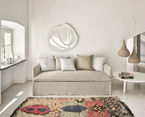 Chambre minimaliste avec un canapé beige, un miroir décoratif, des suspensions tissées et un tapis coloré au sol, apportant une touche d'élégance parisienne. Canape Paris by Atelier Bis