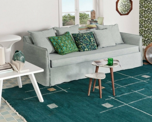 Un salon cosy avec un canapé bleu clair, des coussins à motifs, un tapis vert et des livres empilés dégage un charme parisien. Une table d'appoint blanche complète le look avec une qualité sans effort. Canape Paris by Atelier Bis