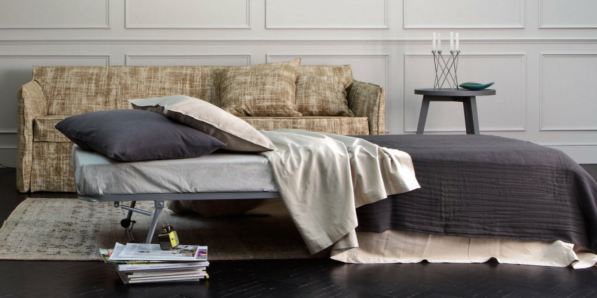 Une chambre cosy avec un canapé convertible en lit, orné d'une literie grise. À côté, une table d'appoint scintille de bougies, tandis que des magazines empilés évoquent le charme de la décoration parisienne et la qualité assurée. Canape Paris by Atelier Bis