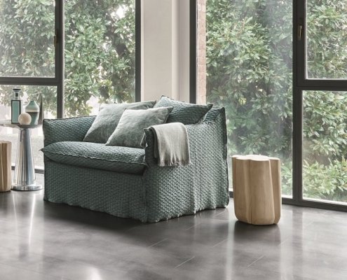 Un fauteuil vert confortable avec des coussins et une couverture se trouve dans une salle d'exposition parisienne, près de grandes fenêtres présentant une verdure luxuriante à l'extérieur. Canape Paris by Atelier Bis