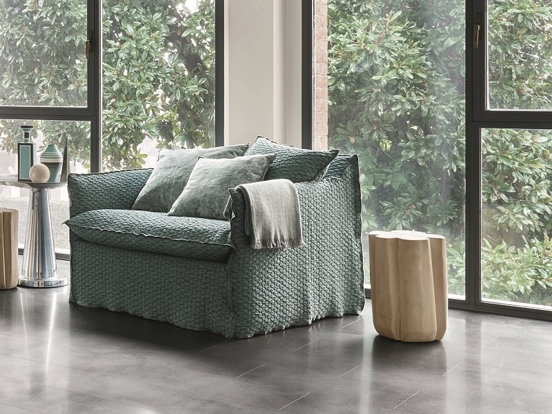 Un fauteuil vert confortable avec des coussins et une couverture se trouve dans une salle d'exposition parisienne, près de grandes fenêtres présentant une verdure luxuriante à l'extérieur. Canape Paris by Atelier Bis