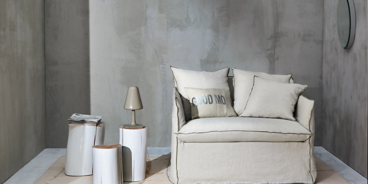 Une salle d'exposition minimaliste comprend un canapé beige clair, des tables d'appoint élégantes, une lampe élégante et des œuvres d'art murales sophistiquées sur un mur texturé gris, le tout présentant une qualité inégalée. Canape Paris by Atelier Bis