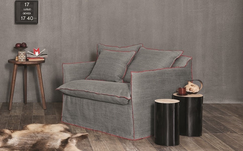 Un fauteuil gris chic avec une bordure rouge, évoquant une touche parisienne, se trouve à côté d'une petite table d'appoint et de deux tabourets en bois sur un plancher en bois avec un tapis en fourrure moelleuse. Canape Paris by Atelier Bis