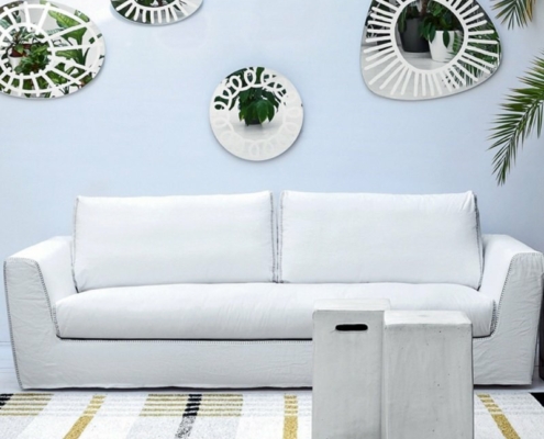 Canapé blanc avec une table basse devant, miroirs décoratifs et plantes ornant le mur au-dessus. Tapis rayé en dessous, comme s'il avait été conçu pour un showroom chic parisien. Canape Paris by Atelier Bis