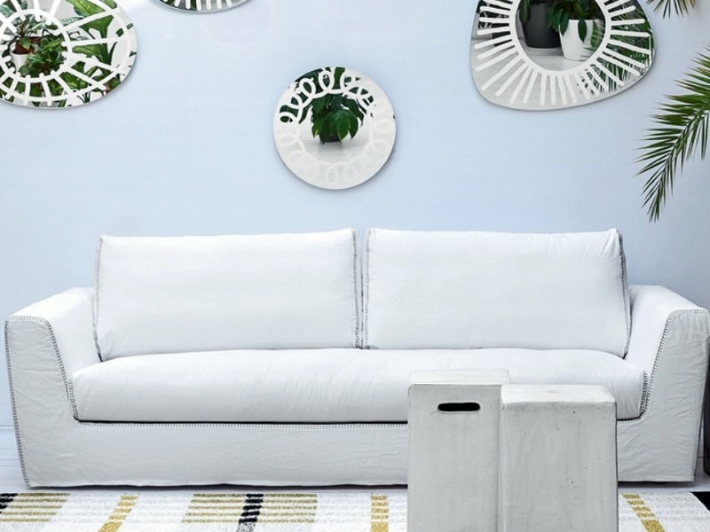 Canapé blanc avec une table basse devant, miroirs décoratifs et plantes ornant le mur au-dessus. Tapis rayé en dessous, comme s'il avait été conçu pour un showroom chic parisien. Canape Paris by Atelier Bis