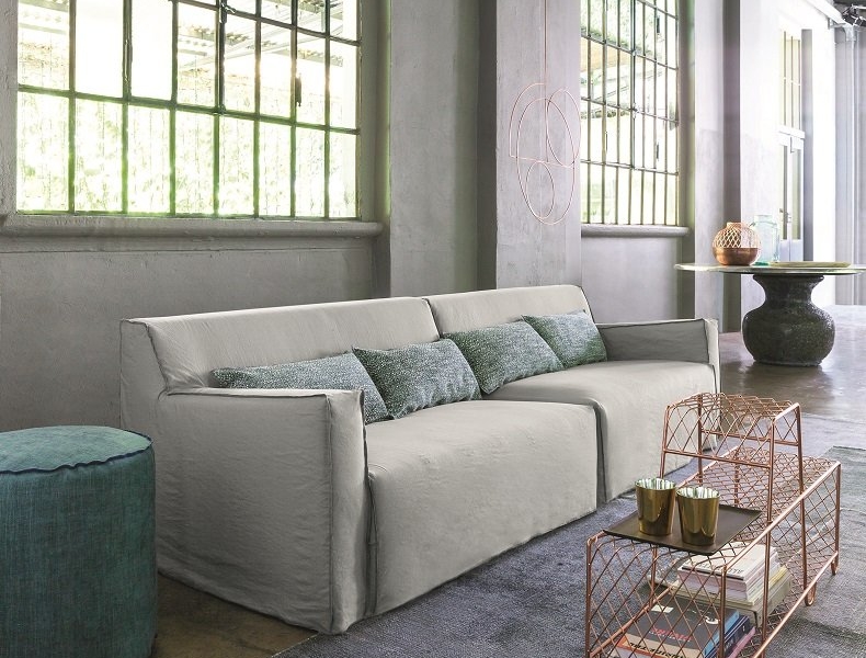 Loft moderne avec un canapé blanc, des coussins bleu canard et une table basse en fil de fer. De hautes fenêtres donnent sur une végétation luxuriante, offrant une touche parisienne qui transforme l'espace en un showroom élégant de haute qualité. Canape Paris by Atelier Bis