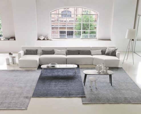 Salon moderne avec un canapé blanc, des tapis gris et beiges, des tables basses et de grandes fenêtres cintrées rappelant un showroom parisien. Canape Paris by Atelier Bis