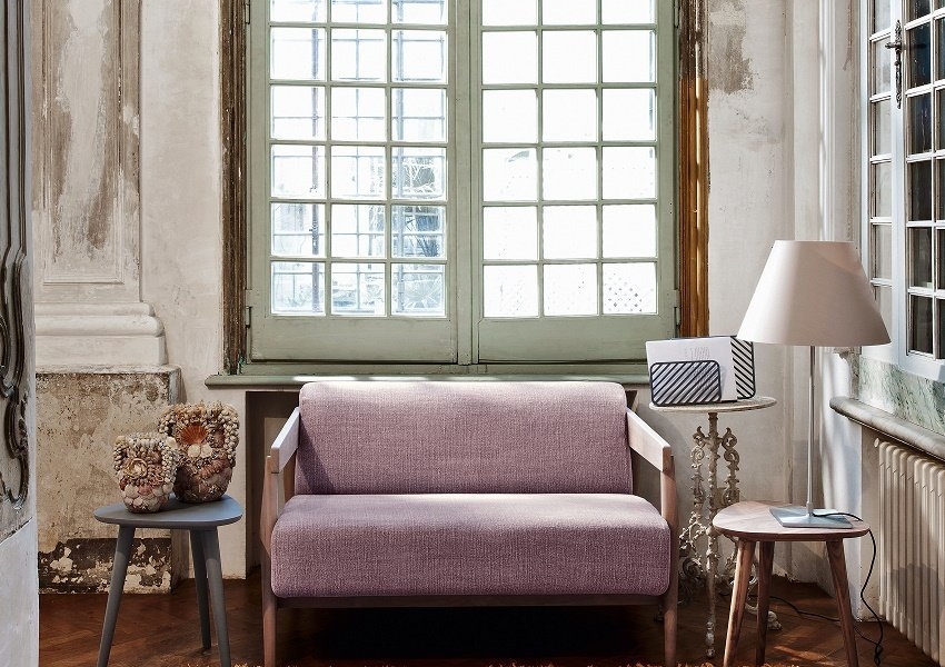 Chambre cosy avec un canapé rose, des tables d'appoint, une lampe et une grande fenêtre rappelant un showroom parisien. Un vase décoratif et un tapis à motifs rehaussent la qualité du décor. Canape Paris by Atelier Bis