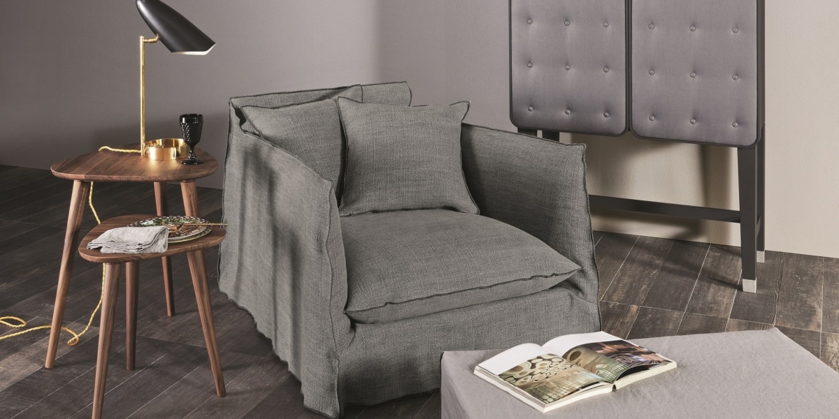 Un fauteuil gris, une table d'appoint en bois avec une lampe et un livre ouvert reposent sur un pouf recouvert dans une pièce moderne et minimaliste qui respire la qualité sophistiquée du design parisien. Canape Paris by Atelier Bis