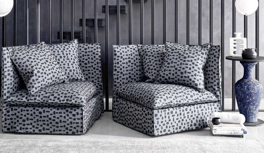 Un salon chic et moderne présente un canapé face à deux chaises grises à motifs, avec un vase bleu ornant la table ronde et des objets décoratifs astucieusement disposés sous l'escalier, évoquant l'élégance de Paris. Canape Paris by Atelier Bis