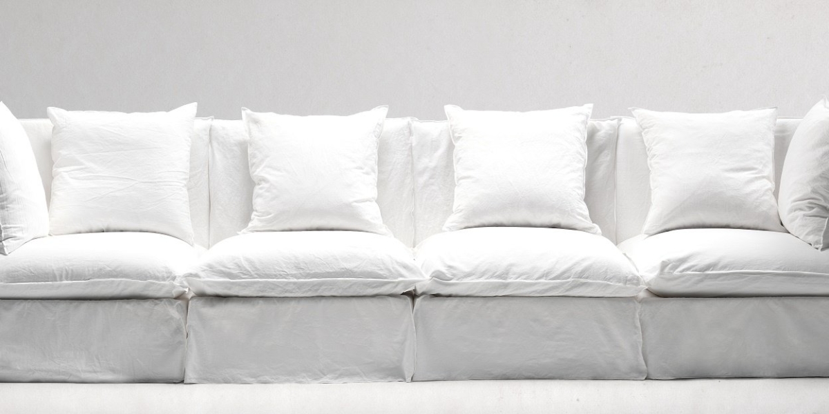 Un long canapé blanc avec plusieurs coussins posés sur un fond blanc uni incarne l'essence de l'élégance parisienne, offrant un mélange harmonieux de style et de confort. Canape Paris by Atelier Bis