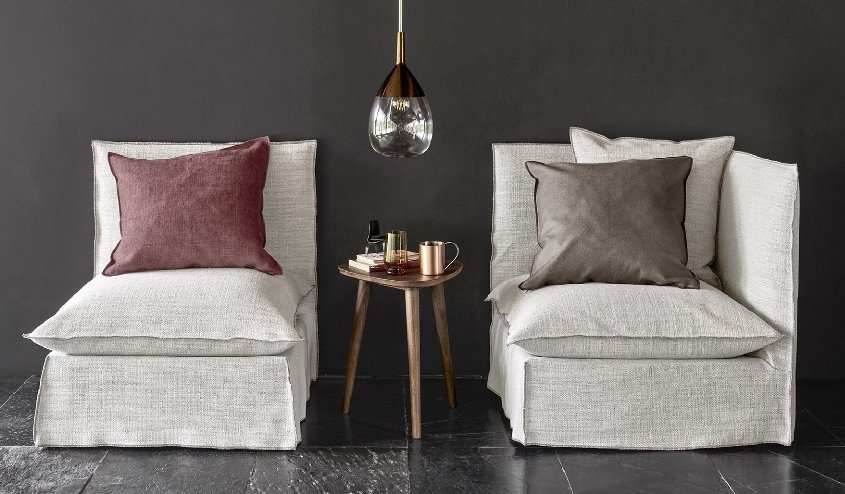 Deux fauteuils beiges avec coussins, une petite table ronde en bois et une suspension contre un mur sombre évoquent le charme de l'élégance parisienne. Canape Paris by Atelier Bis