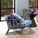 Une chaise en bois gris avec des coussins bleus trône à côté d'une petite table ronde avec une théière, installée à l'intérieur près d'une grande fenêtre. Cette configuration élégante respire le charme parisien, offrant un rapport qualité-prix et style inégalé. Canape Paris by Atelier Bis