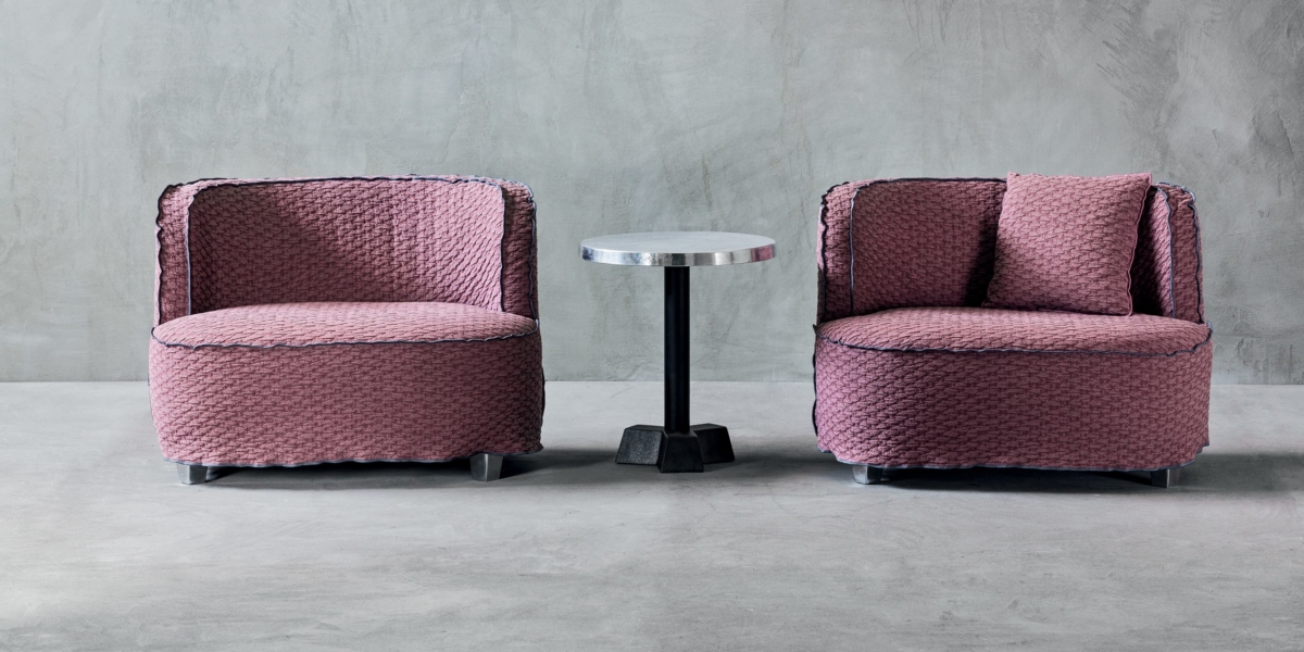 Deux fauteuils texturés violets, rappelant la qualité parisienne, flanquent une petite table d'appoint ronde en métal sur un sol en béton contre un mur gris. Canape Paris by Atelier Bis