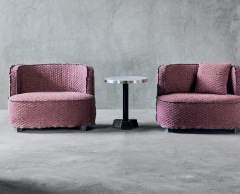 Deux fauteuils texturés violets, rappelant la qualité parisienne, flanquent une petite table d'appoint ronde en métal sur un sol en béton contre un mur gris. Canape Paris by Atelier Bis