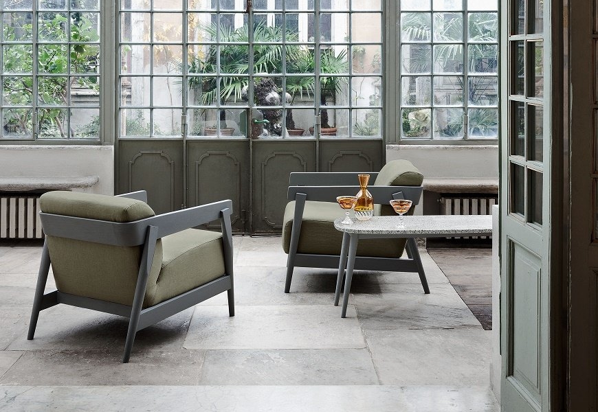 Deux fauteuils verts avec une petite table entre eux, sur laquelle sont posés des verres à vin, sont assis gracieusement dans une pièce ensoleillée avec de grandes fenêtres, offrant une touche de qualité parisienne. Canape Paris by Atelier Bis