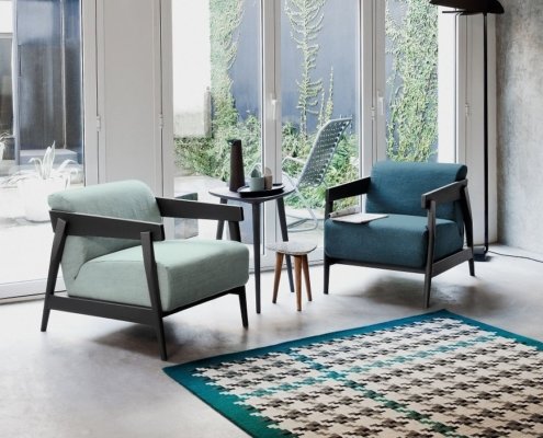 Deux chaises modernes avec une petite table au milieu reposent sur un tapis à motifs, rappelant une configuration de salle d'exposition élégante, dans une pièce ensoleillée avec de grandes fenêtres. Canape Paris by Atelier Bis