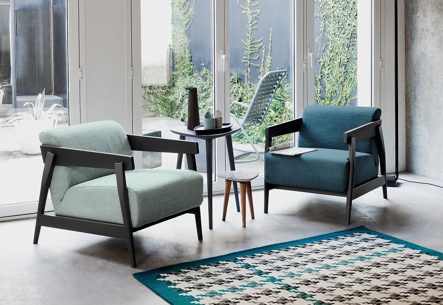 Deux chaises modernes avec une petite table au milieu reposent sur un tapis à motifs, rappelant une configuration de salle d'exposition élégante, dans une pièce ensoleillée avec de grandes fenêtres. Canape Paris by Atelier Bis