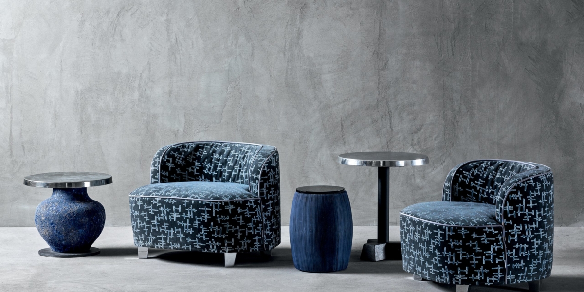 Deux fauteuils à motifs avec de petites tables rondes entre eux créent un coin cosy rappelant un showroom parisien, adossé à un mur et un sol gris texturés. Canape Paris by Atelier Bis