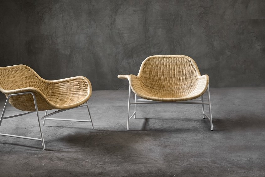 Deux chaises en osier aux pieds en métal reposent sur un sol en béton, adossée à un mur texturé gris foncé, évoquant l'ambiance chic d'un showroom parisien. Canape Paris by Atelier Bis