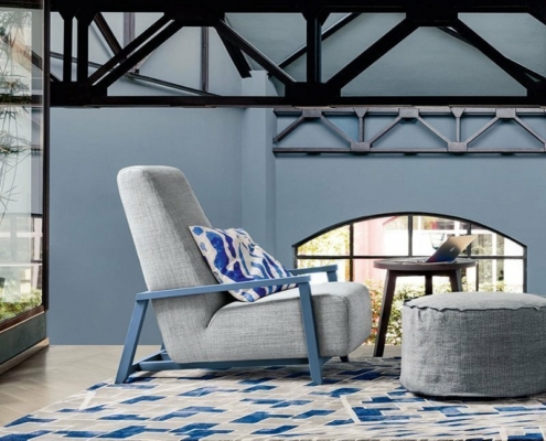 Découvrez un salon moderne avec une chaise grise, un tapis à motifs bleus et un pouf, le tout sur fond de poutres apparentes et de grandes fenêtres. C'est comme entrer dans un showroom chic où chaque détail est soigné à la perfection. Canape Paris by Atelier Bis