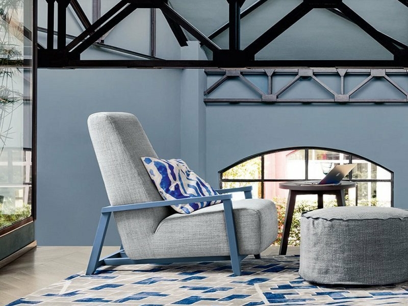 Découvrez un salon moderne avec une chaise grise, un tapis à motifs bleus et un pouf, le tout sur fond de poutres apparentes et de grandes fenêtres. C'est comme entrer dans un showroom chic où chaque détail est soigné à la perfection. Canape Paris by Atelier Bis