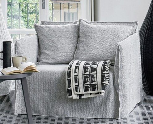 Un fauteuil gris avec une housse texturée, deux coussins et une couverture à motifs noirs et blancs évoquent l'élégance chaleureuse d'un salon parisien. Canape Paris by Atelier Bis