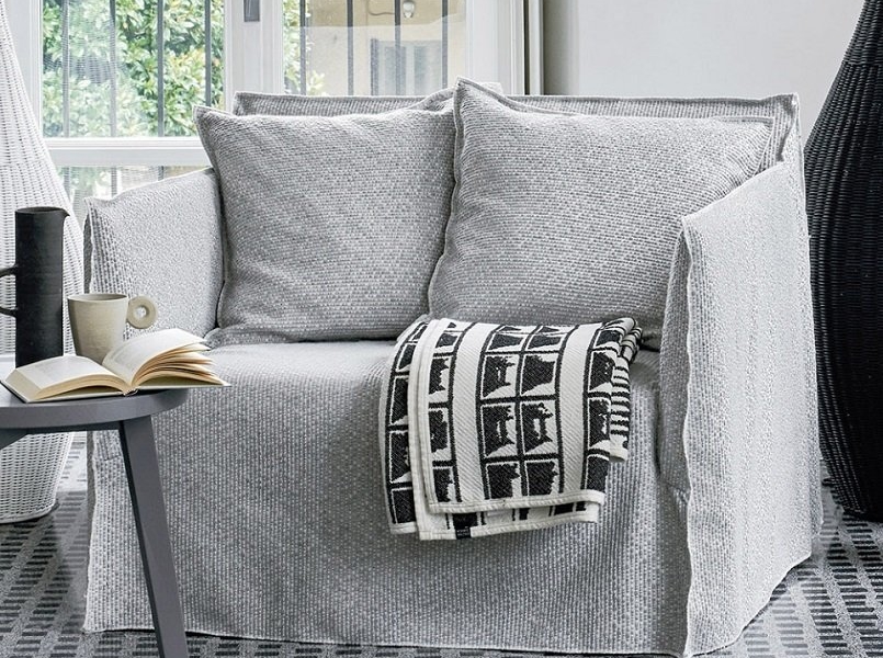 Un fauteuil gris avec une housse texturée, deux coussins et une couverture à motifs noirs et blancs évoquent l'élégance chaleureuse d'un salon parisien. Canape Paris by Atelier Bis