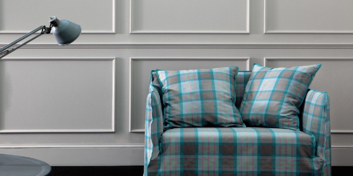 Un fauteuil recouvert de tissu écossais avec des coussins assortis trône élégamment à côté d'une lampe et d'une table grises contre un mur lambrissé, rappelant la qualité et le charme parisiens. Canape Paris by Atelier Bis