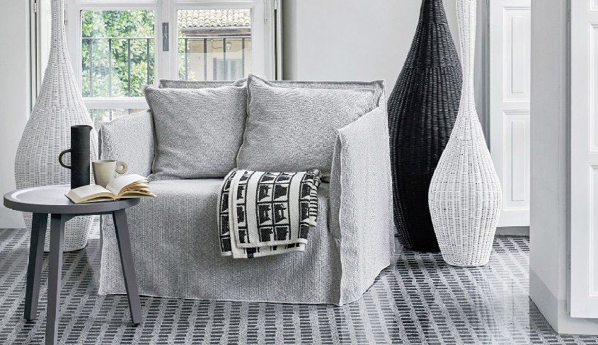 Fauteuil avec revêtement gris et couverture à motifs, installé dans le coin d'un showroom parisien. Une table ronde à proximité accueille un livre, une tasse et deux grands vases tressés, créant un espace invitant à la détente. Canape Paris by Atelier Bis