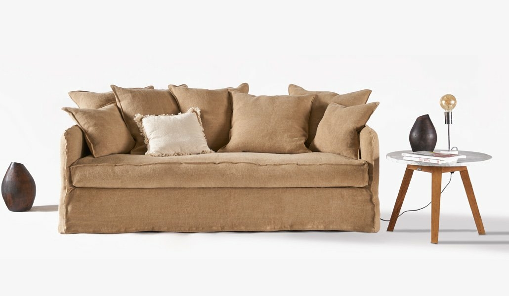 Un canapé marron orné de plusieurs coussins trône élégamment à côté d'une petite table. Au-dessus, une lampe diffuse une lumière chaleureuse et à côté, un vase ajoute de l'élégance. Cette configuration sereine rappelle une vitrine immaculée sur un fond sobre. Canape Paris by Atelier Bis