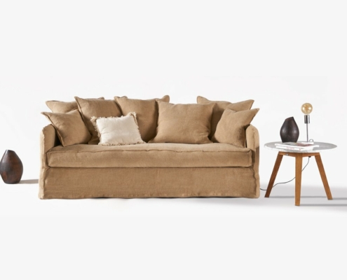 Un canapé marron orné de plusieurs coussins trône élégamment à côté d'une petite table. Au-dessus, une lampe diffuse une lumière chaleureuse et à côté, un vase ajoute de l'élégance. Cette configuration sereine rappelle une vitrine immaculée sur un fond sobre. Canape Paris by Atelier Bis