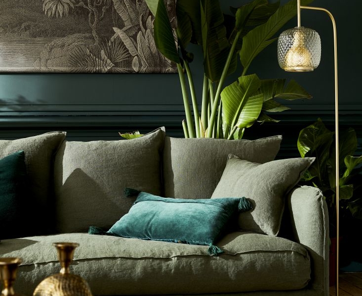 Un salon chaleureux avec canapé vert, entouré de plantes tropicales et agrémenté d'œuvres d'art murales sur le thème de la nature, éclairé par un lampadaire élégant. Profitez du mélange parfait de qualité et de style dans cet espace chaleureux. Canape Paris by Atelier Bis