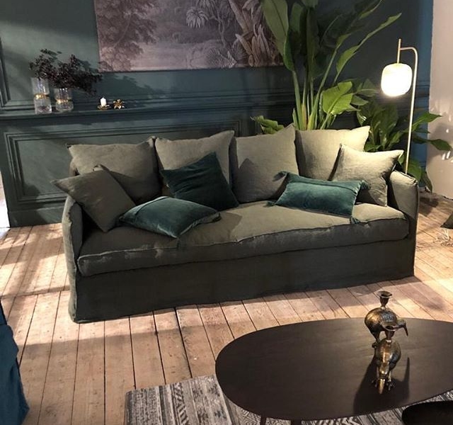 Un salon d'inspiration parisienne dispose d'un canapé vert orné de plusieurs coussins, complété par un décor végétal et une table basse en bois posée sur un tapis texturé. Canape Paris by Atelier Bis