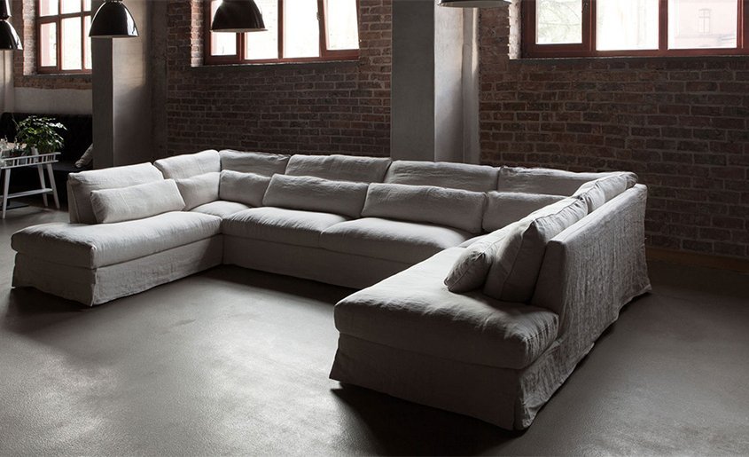 Un salon spacieux ressemblant à une salle d'exposition parisienne dispose d'un grand canapé sectionnel blanc et de murs en briques, éclairés par de larges fenêtres et des suspensions. Canape Paris by Atelier Bis