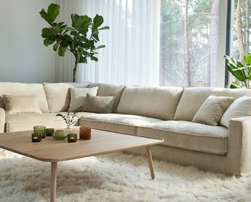 Salon moderne avec un grand canapé beige, une table basse en bois, des plantes vertes et un piano ; de grandes fenêtres en arrière-plan créent une atmosphère de salle d'exposition. Canape Paris by Atelier Bis