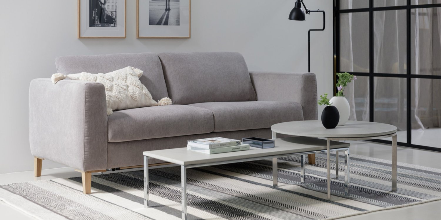 Entrez dans ce salon moderne, rappelant un showroom parisien chic, avec un canapé gris, deux tables basses rondes et une lampe noire épurée. Des photos encadrées ornent le mur blanc immaculé, ajoutant une touche élégante à ce décor sophistiqué. Canape Paris by Atelier Bis