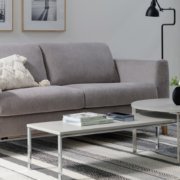 Entrez dans ce salon moderne, rappelant un showroom parisien chic, avec un canapé gris, deux tables basses rondes et une lampe noire épurée. Des photos encadrées ornent le mur blanc immaculé, ajoutant une touche élégante à ce décor sophistiqué. Canape Paris by Atelier Bis