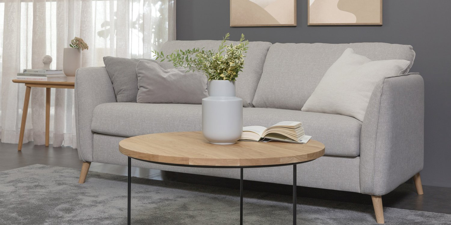 Canapé gris avec coussins, table basse ronde en bois avec vase et livre, art abstrait et table d'appoint dans un salon. Canape Paris by Atelier Bis