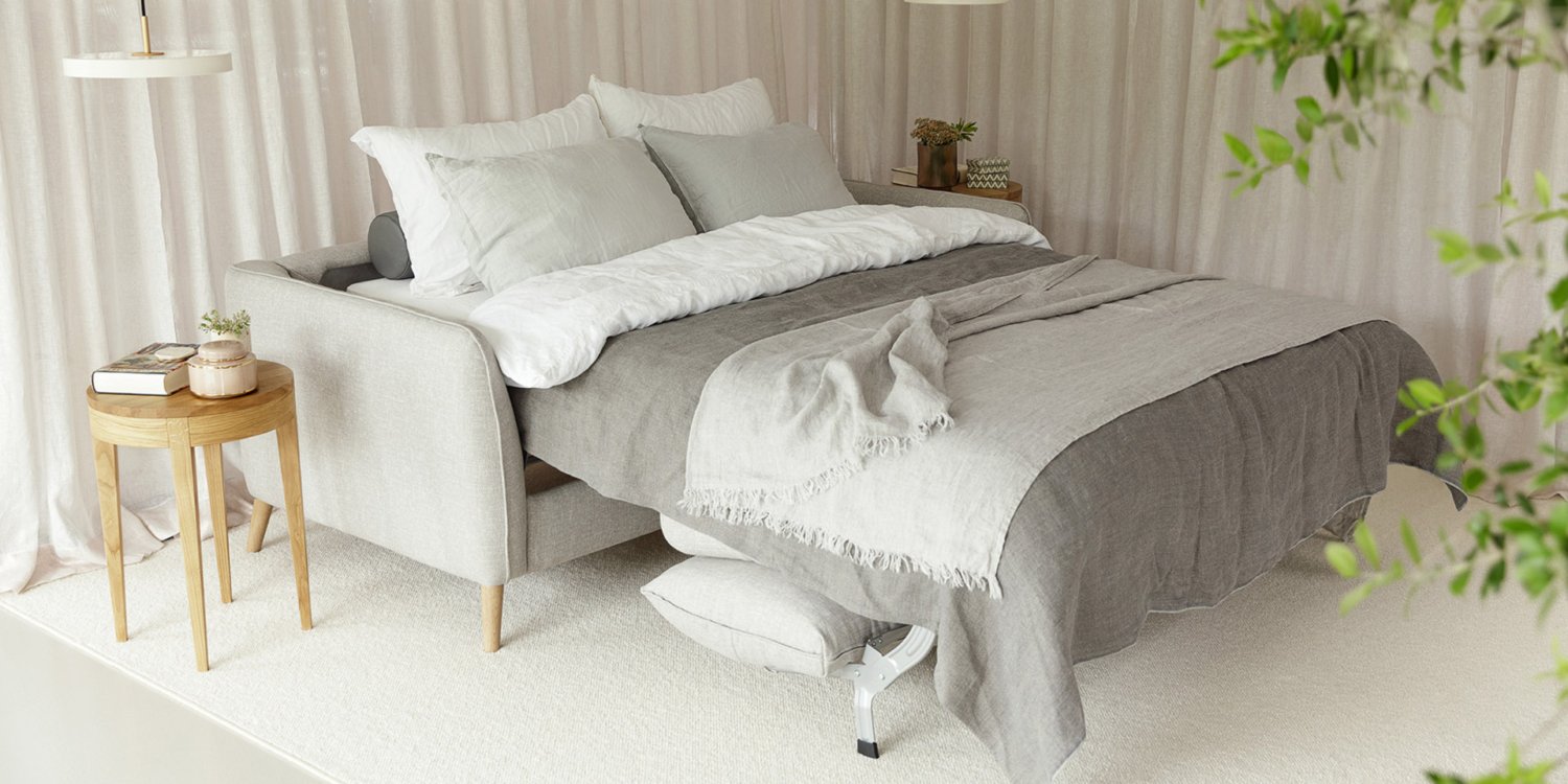 Un lit douillet avec une literie grise et blanche, entouré de rideaux légers rappelant une journée parisienne, et une petite table d'appoint en bois ornée de livres et de plantes. Canape Paris by Atelier Bis