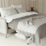 Un lit douillet avec une literie grise et blanche, entouré de rideaux légers rappelant une journée parisienne, et une petite table d'appoint en bois ornée de livres et de plantes. Canape Paris by Atelier Bis