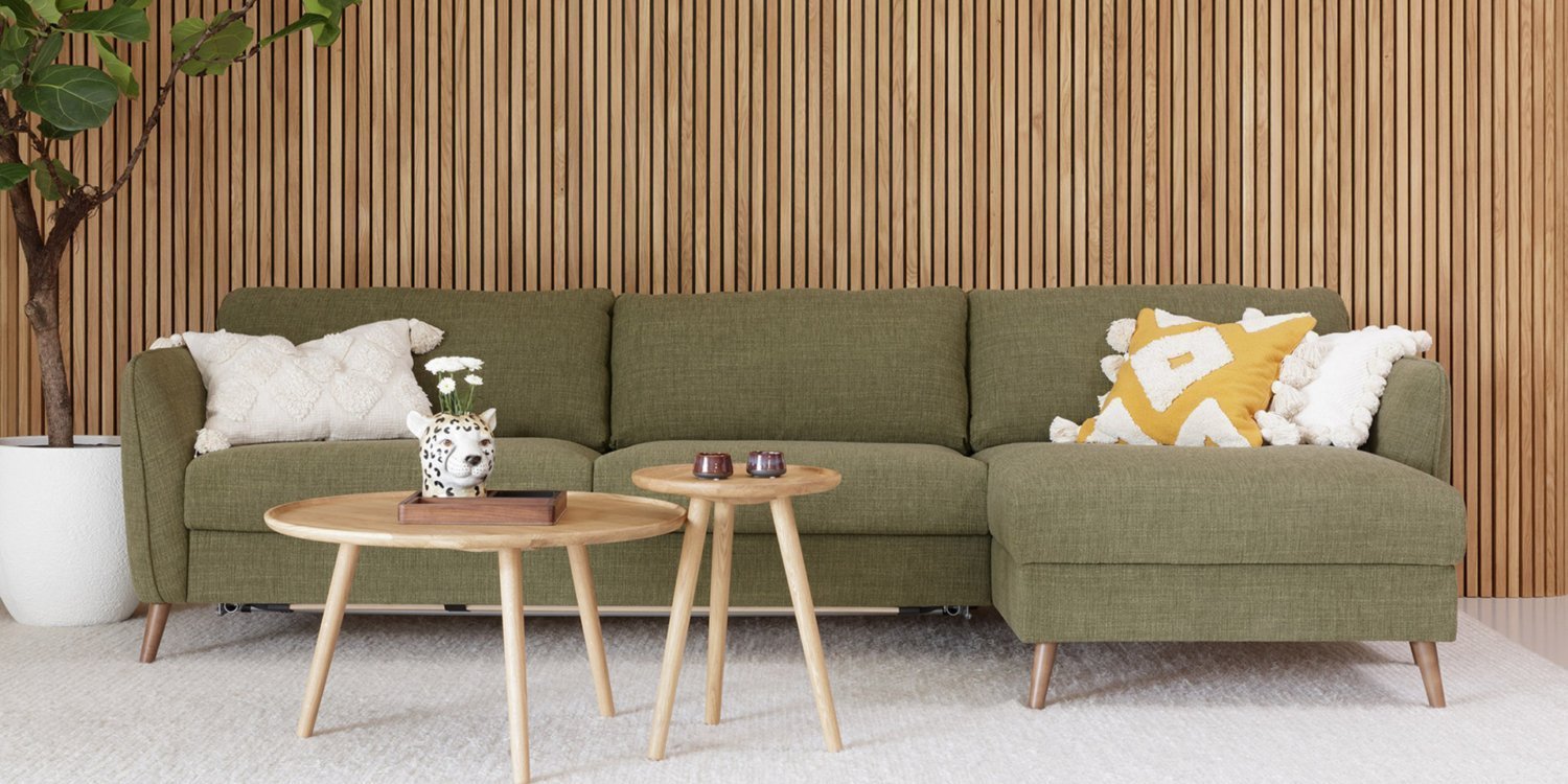 Un salon moderne dispose d'un élégant canapé vert, de deux tables en bois, de coussins décoratifs et d'une plante en pot sur un élégant panneau de bois, rappelant l'ambiance sophistiquée d'une salle d'exposition. Canape Paris by Atelier Bis